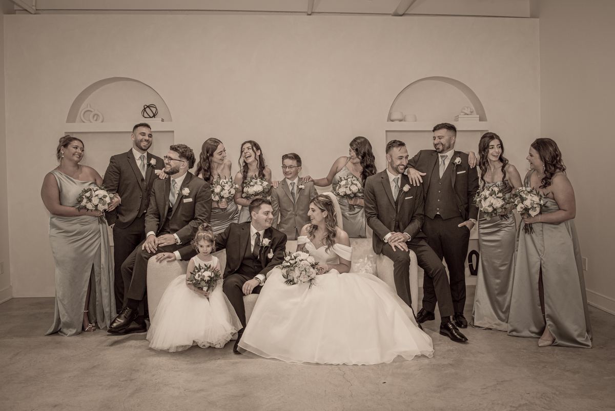 Bride and groom sit center in a warm, sepia-toned group portrait surrounded by smiling wedding party members.