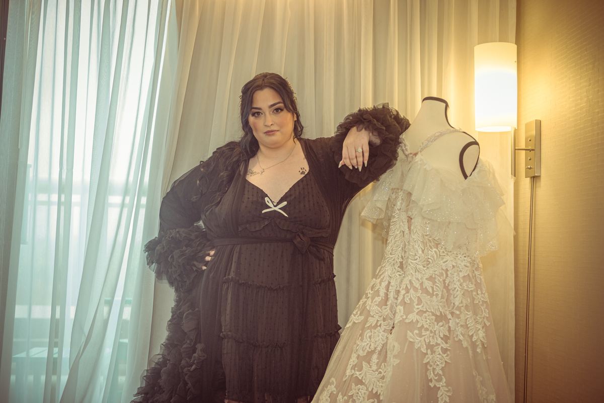 A woman in a dark ruffled dress stands beside a wedding gown on a mannequin, hand resting on its shoulder.