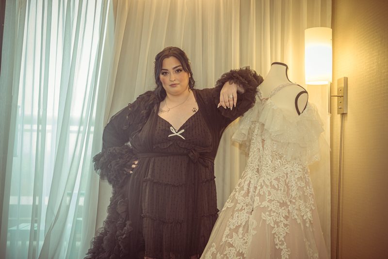 A woman in a dark ruffled dress stands beside a wedding gown on a mannequin, hand resting on its shoulder.