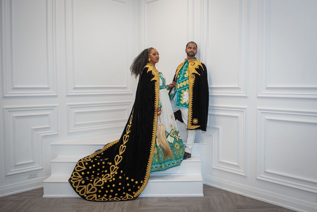 A couple in ornate black and gold ceremonial attire stand on white stairs, holding hands in an intimate pose.