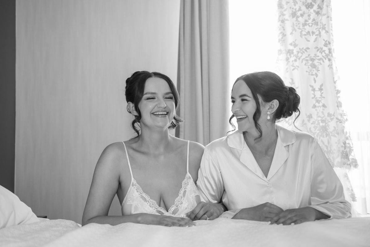 Two women laugh joyfully while seated on a bed in soft, natural light filtering through sheer curtains.