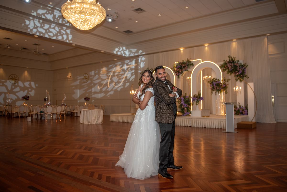 A smiling bride and groom embrace on a dance floor under soft lighting with an illuminated floral arch behind them.