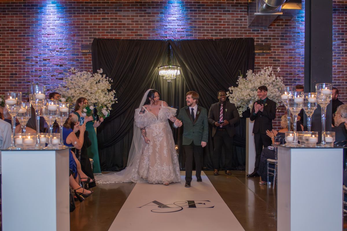 Bride and groom walk down candlelit aisle toward altar, surrounded by applauding guests and floral arrangements.