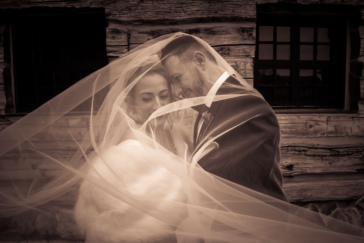 A newlywed couple in traditional attire gazes affectionately at each other near a rustic wooden cabin.