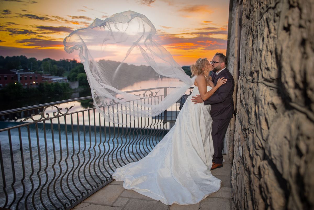 A bride and groom kiss on a stone balcony at sunset, their flowing veil catching the wind over a river.
