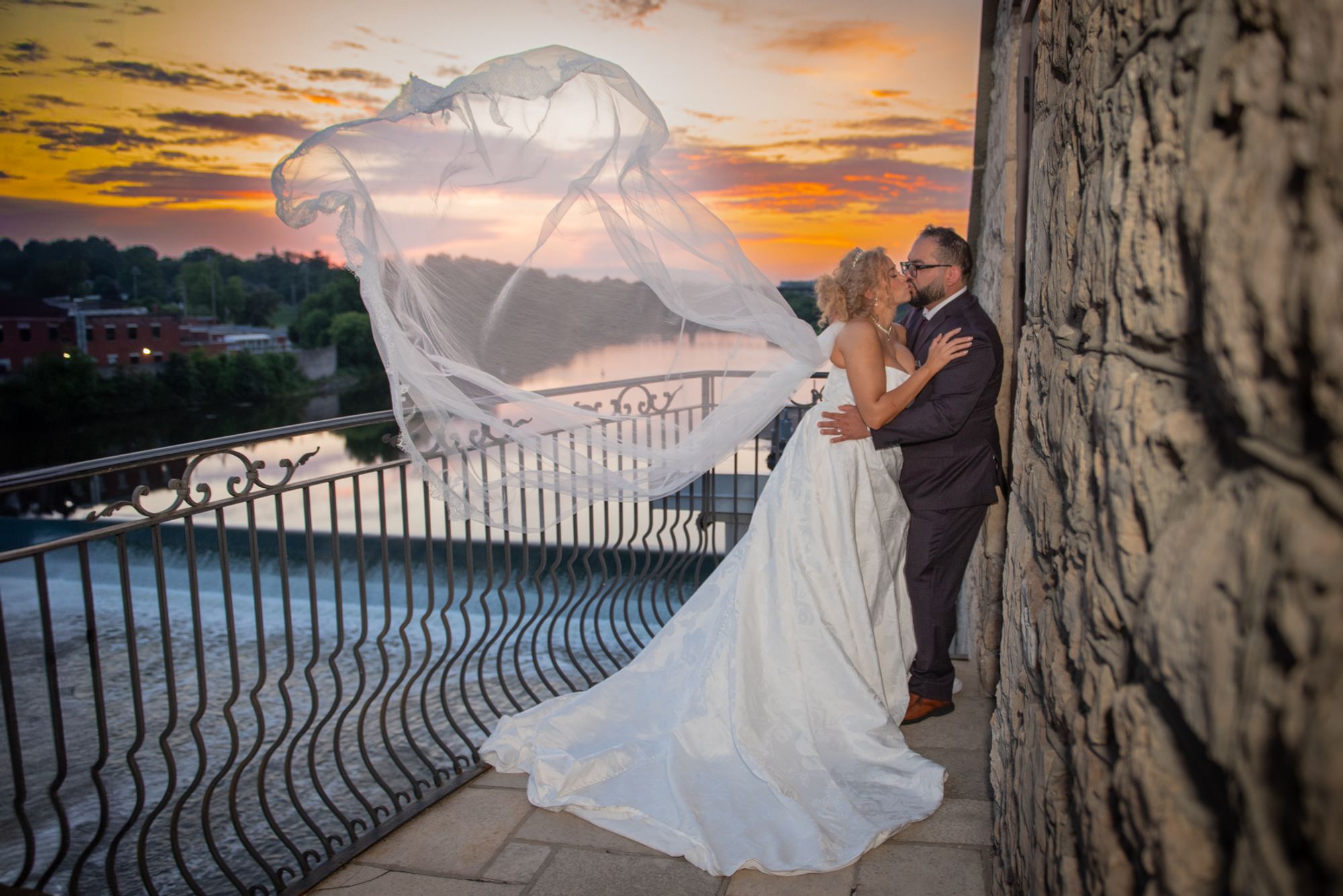 A bride and groom kiss on a stone balcony at sunset, their flowing veil catching the wind over a river.