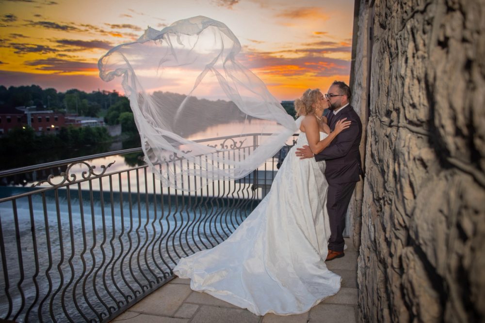 A bride and groom kiss on a stone balcony at sunset, their flowing veil catching the wind over a river.