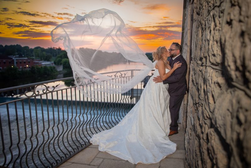 A bride and groom kiss on a stone balcony at sunset, their flowing veil catching the wind over a river.