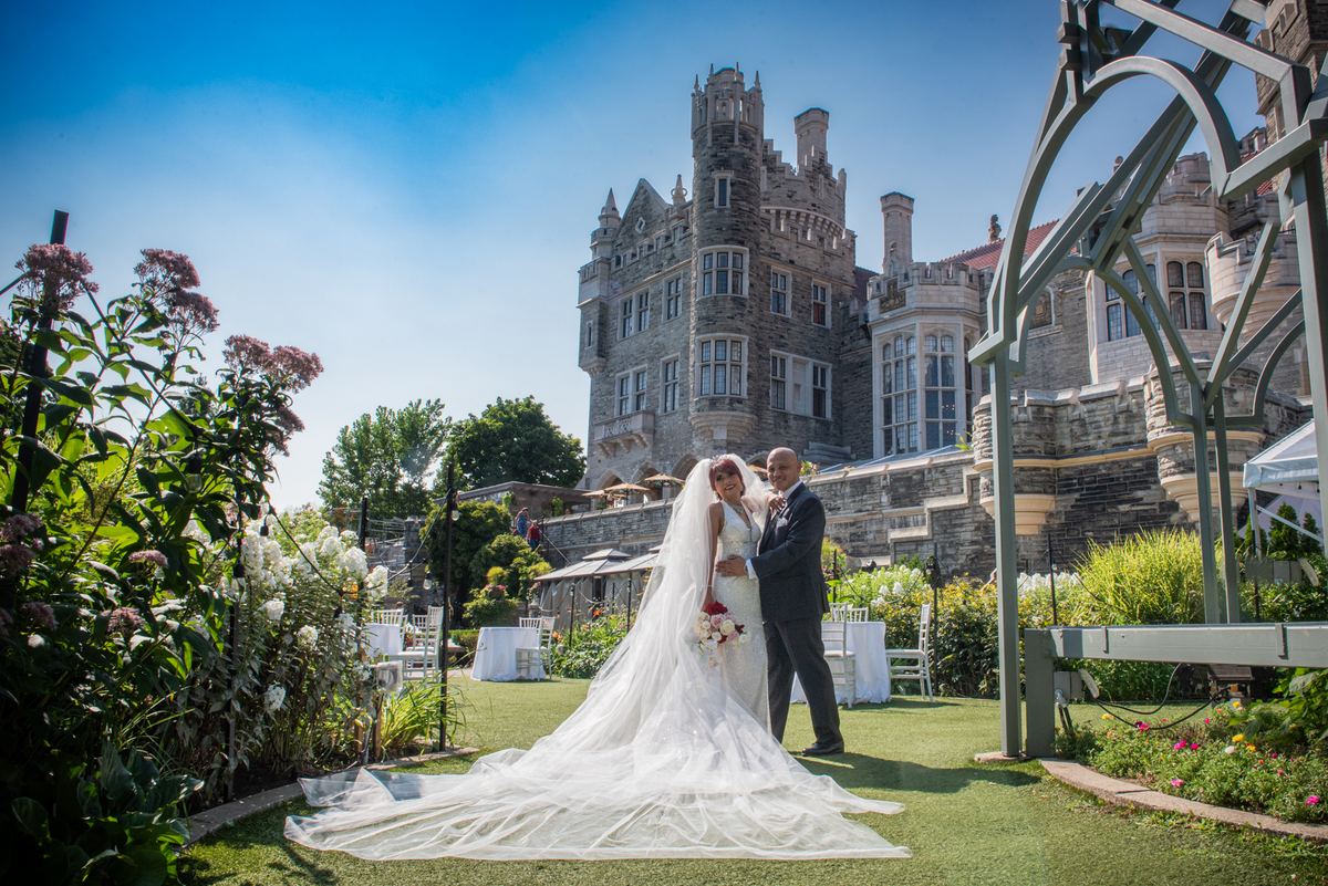 A smiling bride and groom embrace on a sunlit lawn surrounded by lush greenery and a grand stone building.
