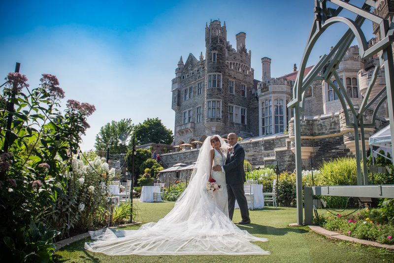 A smiling bride and groom embrace on a sunlit lawn surrounded by lush greenery and a grand stone building.