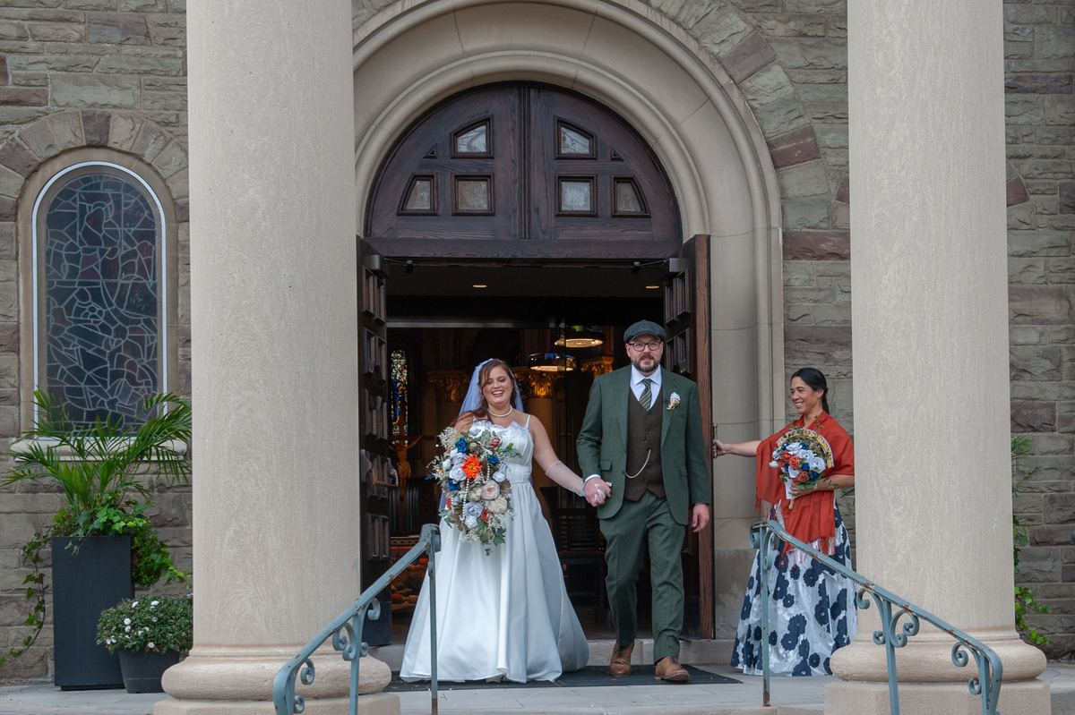 A smiling bride and groom exit a stone church with a bridesmaid beside them, bathed in soft daylight.