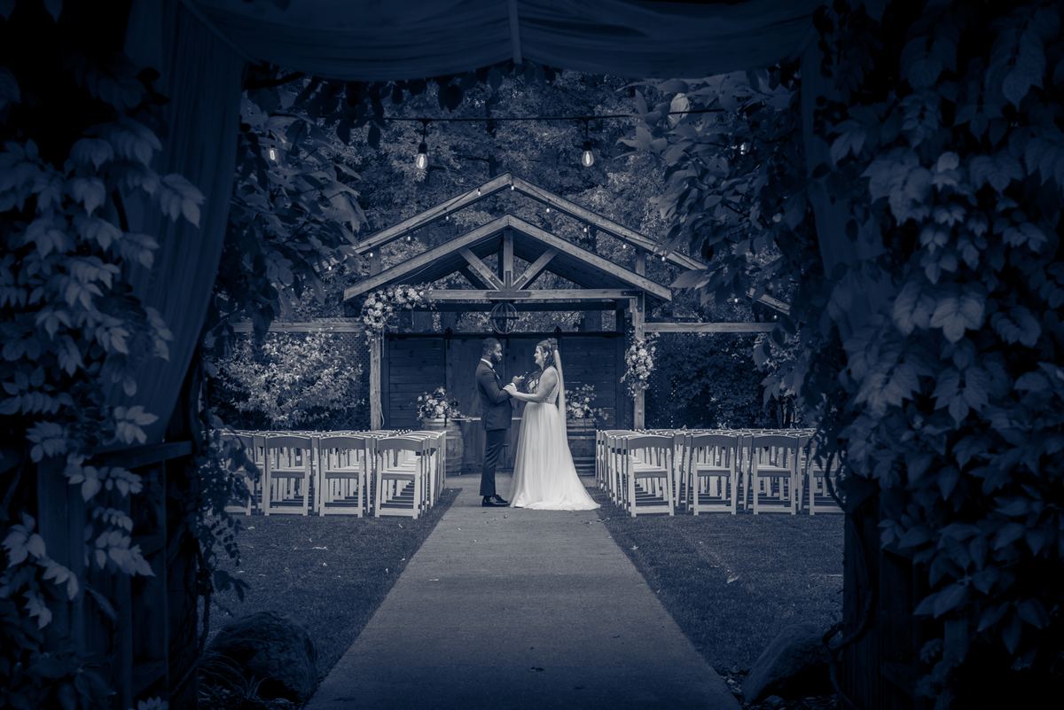 A bride and groom stand hand-in-hand at the center of a rustic outdoor aisle under hanging string lights.