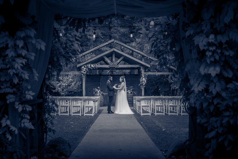 A bride and groom stand hand-in-hand at the center of a rustic outdoor aisle under hanging string lights.