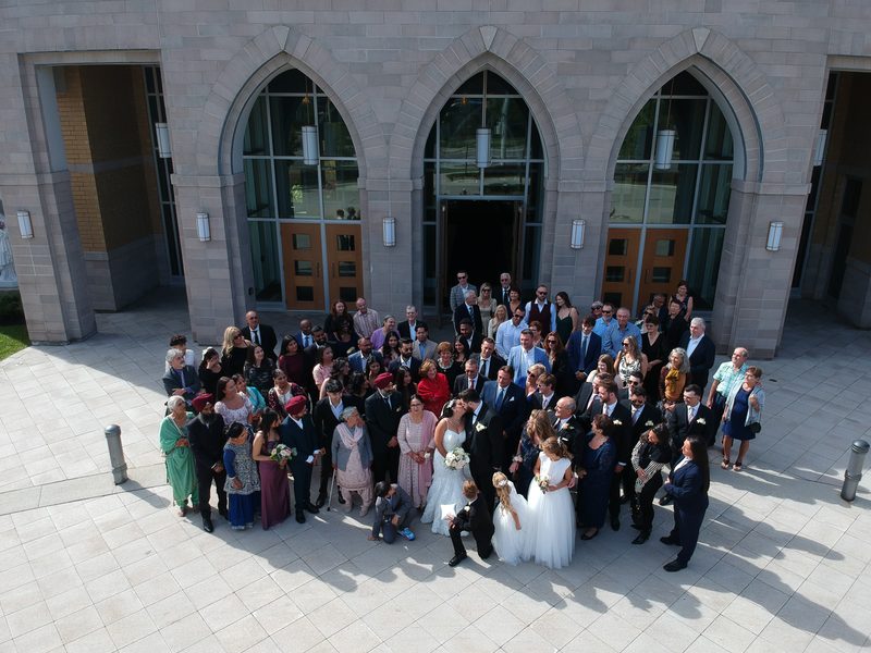 A wedding party in formal attire poses outside a modern stone church with arched glass doors under clear skies.