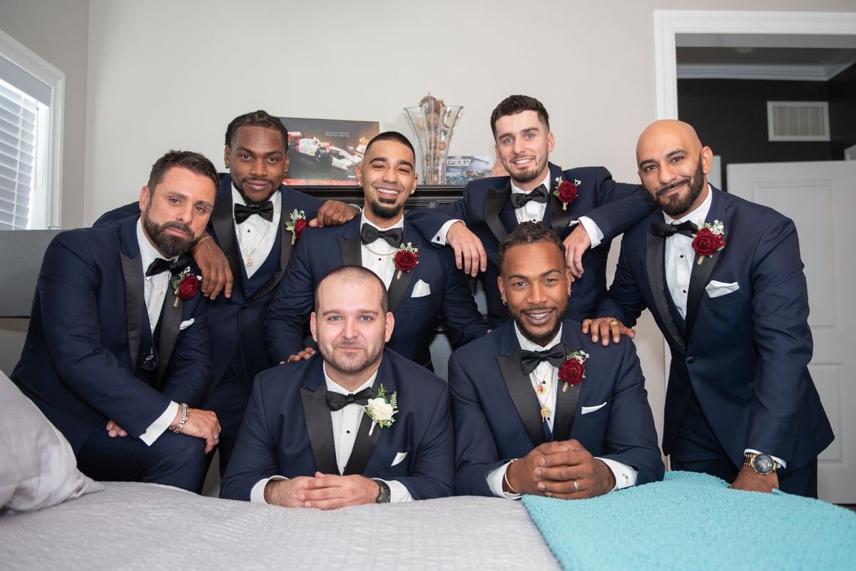 Seven groomsmen in navy tuxedos and red boutonnieres pose together in a bedroom, some standing, some on a bed.