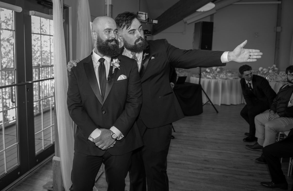 Groom with beard and boutonnière stands beside groomsmate gesturing towards ceremony area, guests in background.
