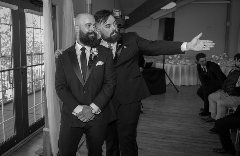 Groom with beard and boutonnière stands beside groomsmate gesturing towards ceremony area, guests in background.