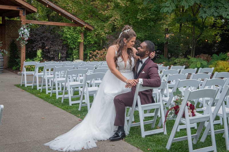 Bride kisses groom's cheek under wooden arbor adorned with flowers and greenery, natural light filtering through trees.