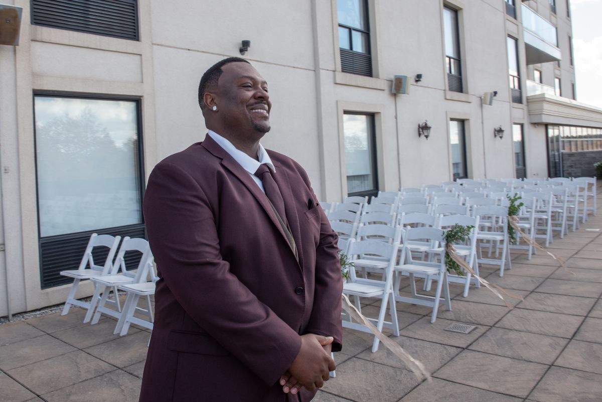 A smiling groom in a maroon suit stands beside white folding chairs arranged for an outdoor wedding ceremony.