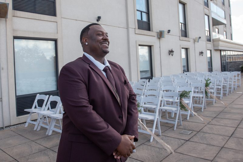 A smiling groom in a maroon suit stands beside white folding chairs arranged for an outdoor wedding ceremony.