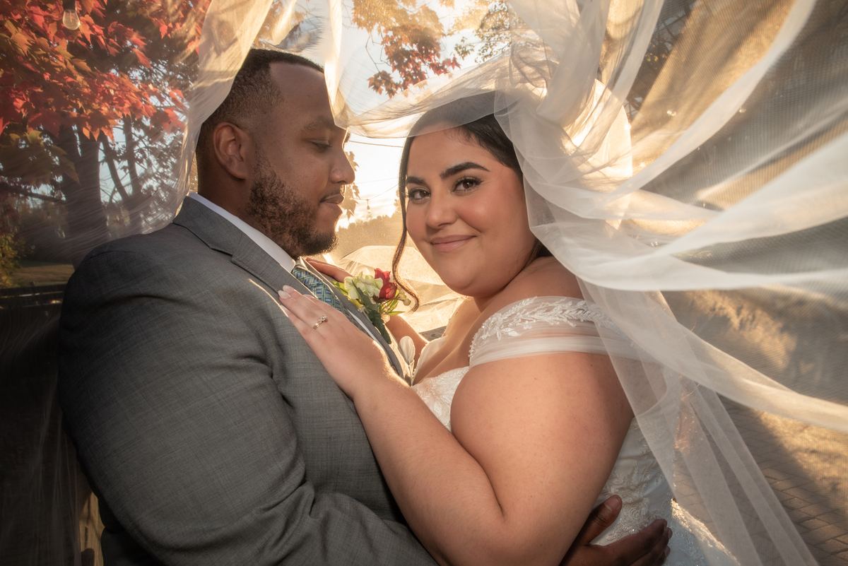 A newlywed couple embraces outdoors as golden sunlight filters through autumn leaves, illuminating the bride's veil.