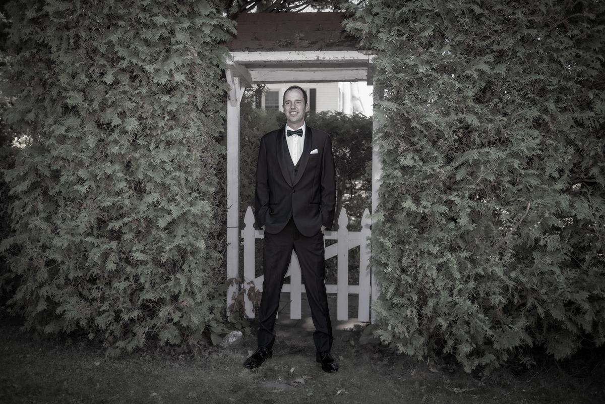 A man in a formal black tuxedo with a bow tie stands smiling in front of a white picket gate surrounded by evergreen hedges.