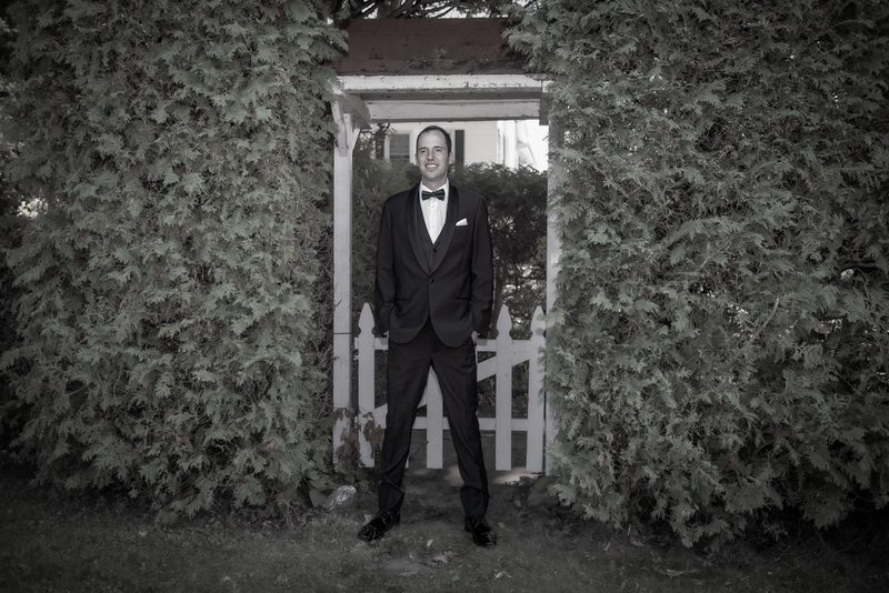 A man in a formal black tuxedo with a bow tie stands smiling in front of a white picket gate surrounded by evergreen hedges.