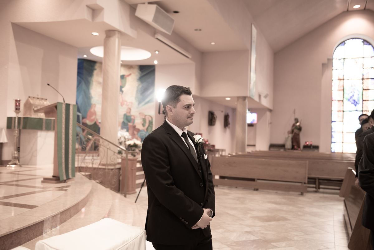 A groom in a black suit stands inside a modern church with stained-glass light and religious iconography.