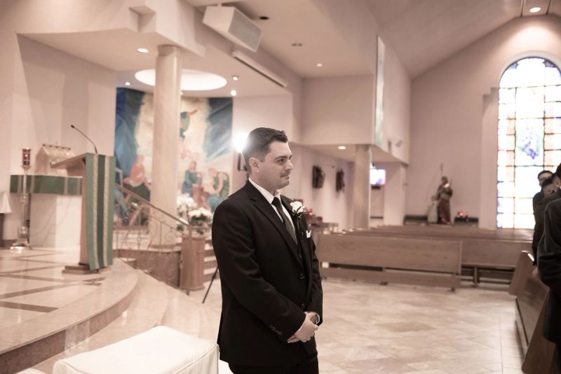 A groom in a black suit stands inside a modern church with stained-glass light and religious iconography.