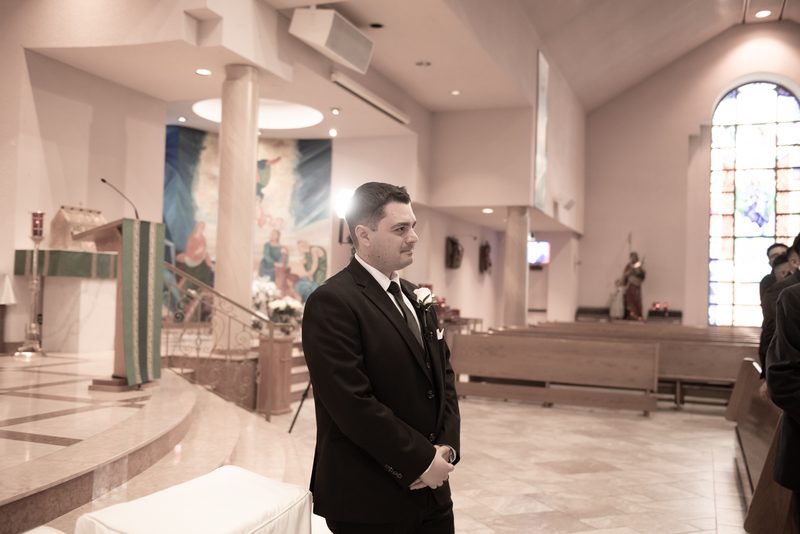 A groom in a black suit stands inside a modern church with stained-glass light and religious iconography.