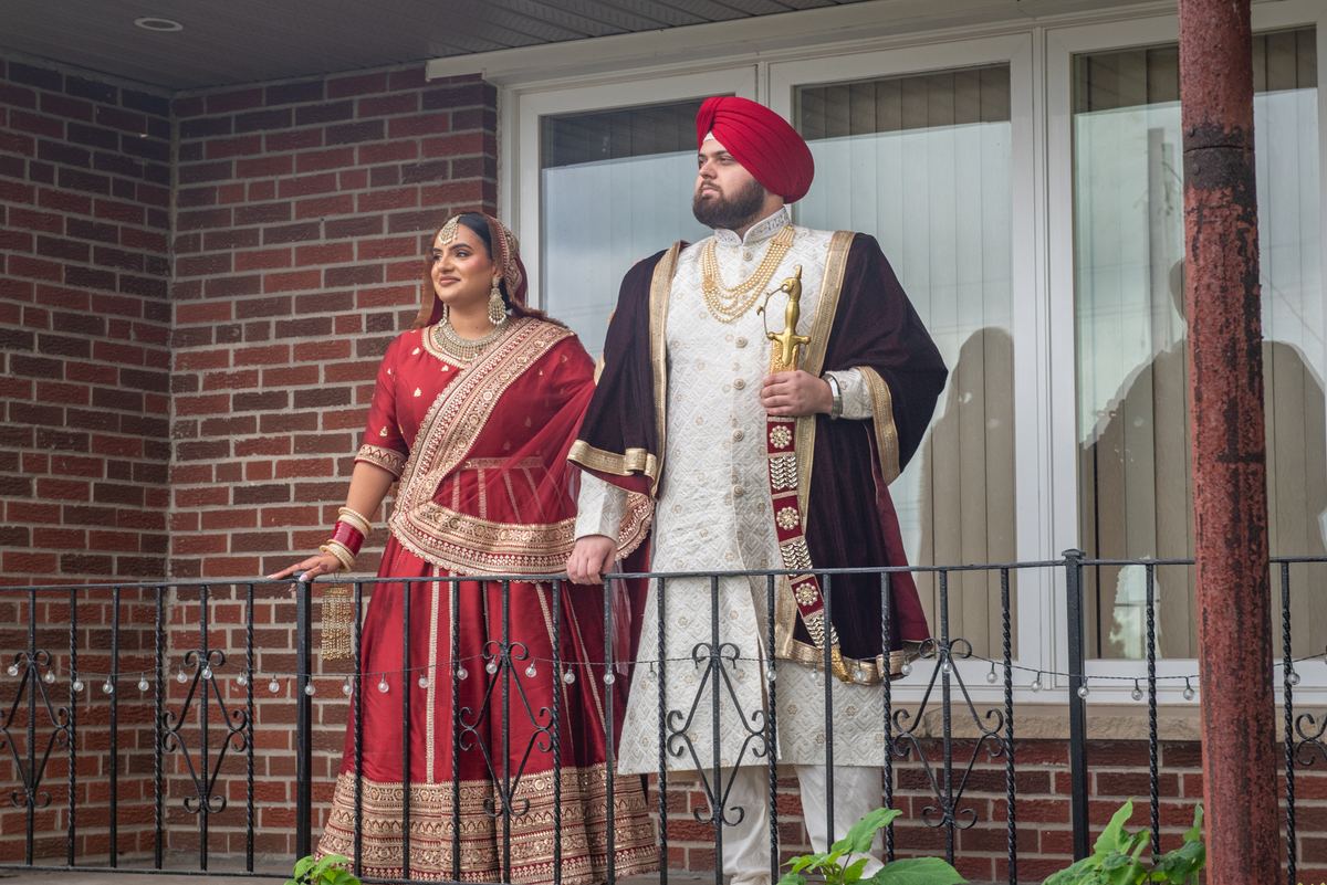A bride and groom in traditional South Asian attire stand on a balcony, gazing outward at a brick wall.
