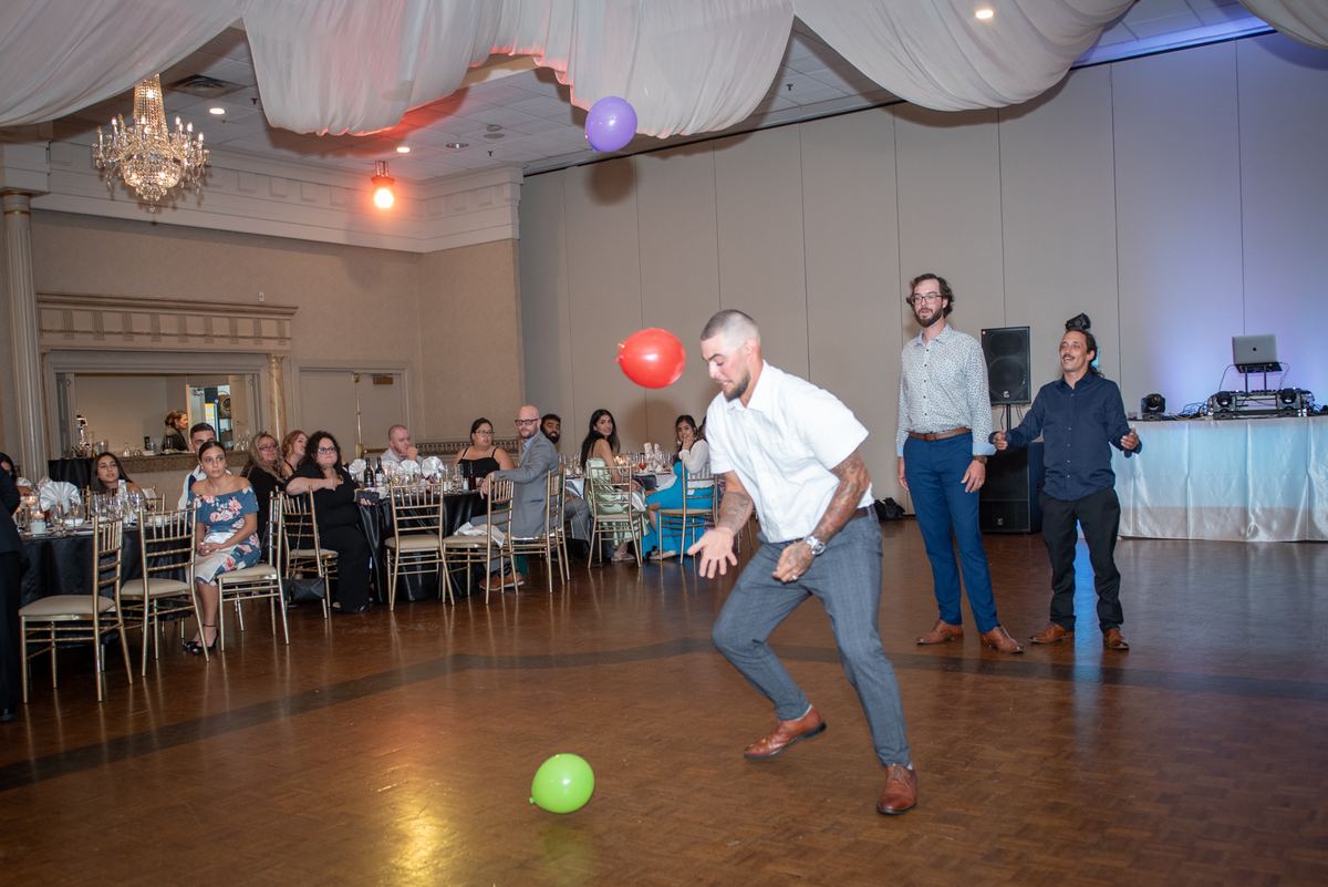 A man leans forward to pop colorful balloons while guests watch amusedly and two men gesture nearby in a warmly lit ballroom.