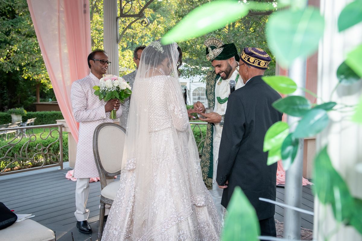 Bride in white gown and veil exchanges rings with groom in green and gold under dappled sunlight, surrounded by guests.