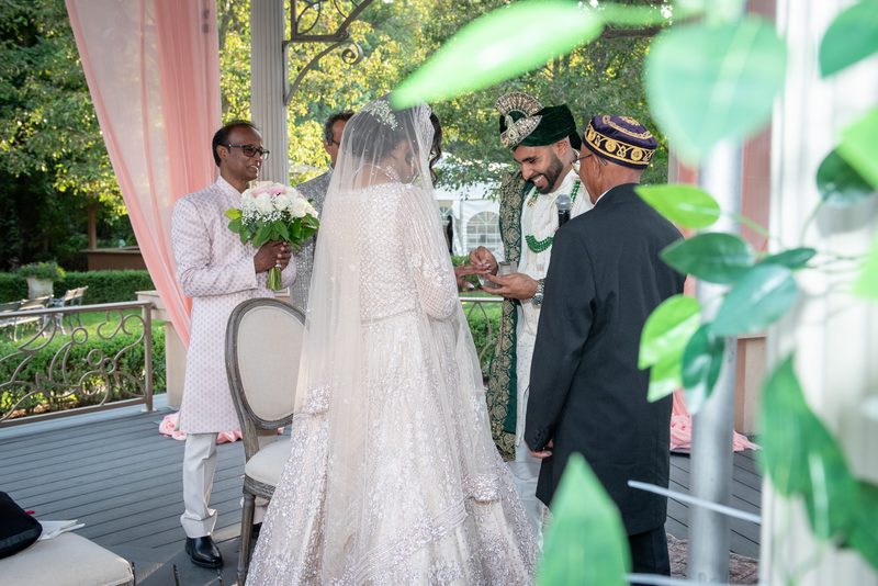 Bride in white gown and veil exchanges rings with groom in green and gold under dappled sunlight, surrounded by guests.