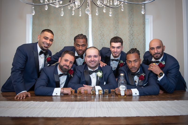 Groom and groomsmen in navy tuxedos gather around a wooden table with shot glasses and a bottle of liquor.