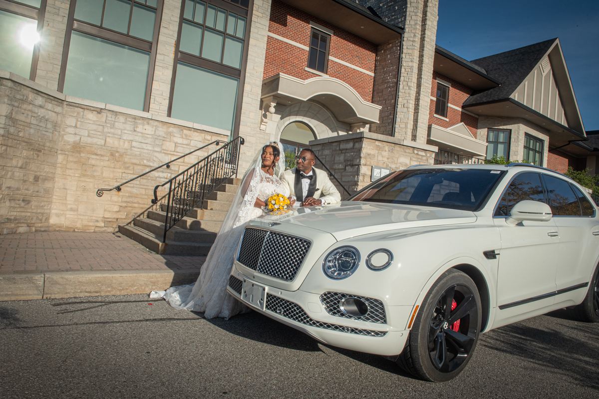 A bride and groom stand beside a white Bentley SUV outside a brick building, holding hands and smiling.