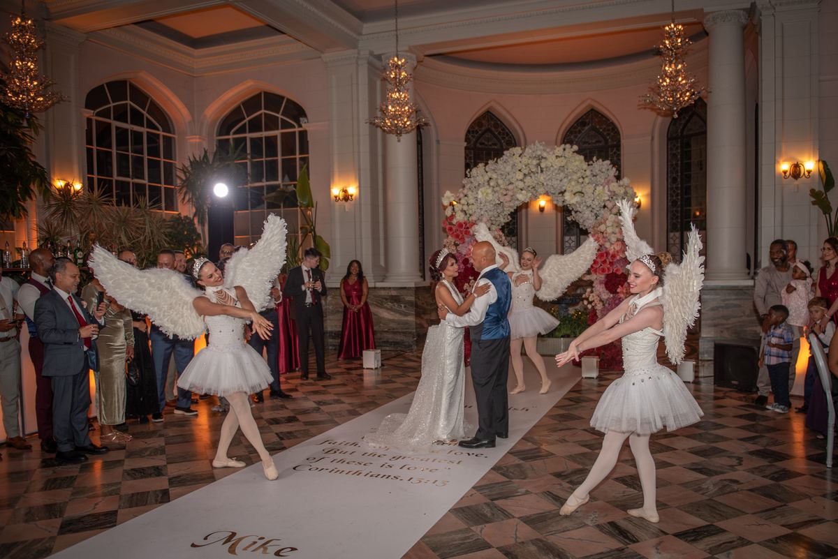 A bride and groom dance tenderly in a ballroom as angelic dancers perform beside them, surrounded by guests.