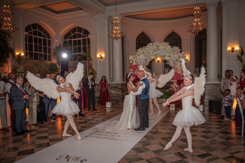 A bride and groom dance tenderly in a ballroom as angelic dancers perform beside them, surrounded by guests.