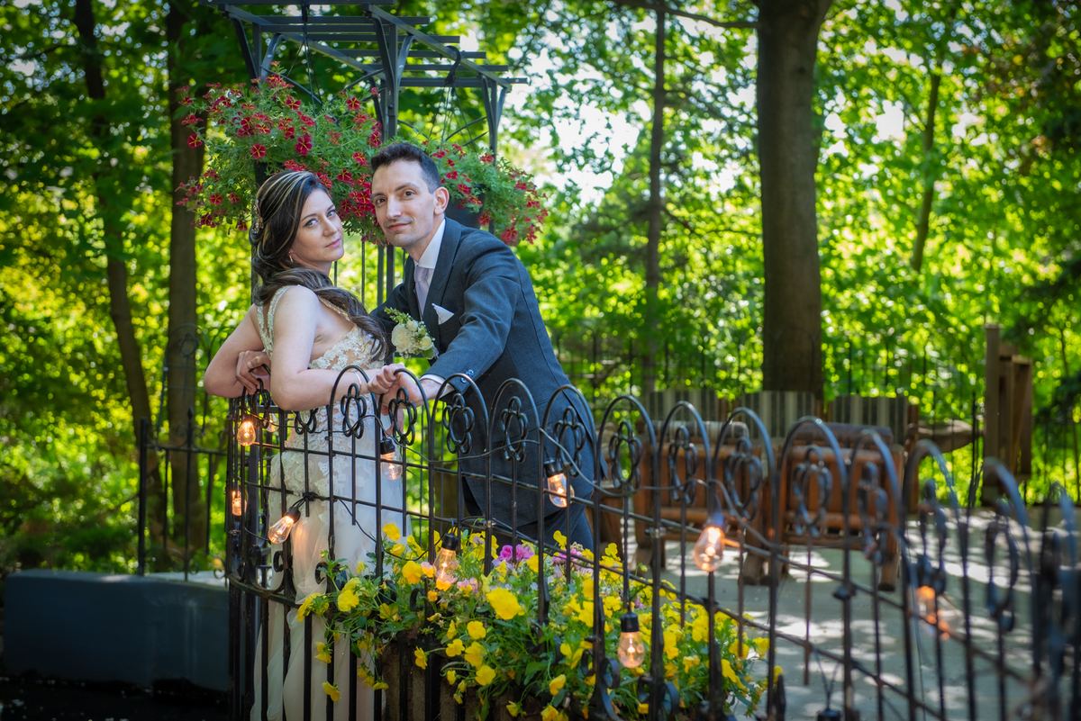 A bride and groom stand beside an ornate fence in a sunlit garden, smiling at the camera as string lights hang above them.