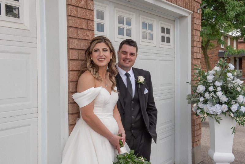 Vittoria and Vince smile warmly for the camera, standing beside a white garage door framed by brickwork.