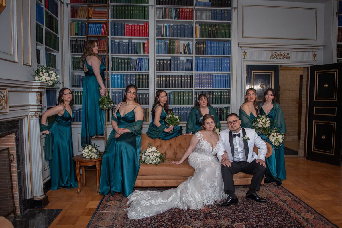 Emily and Ramon are seated on a tufted sofa in a grand library, with bridesmaids in teal dresses surrounding them.
