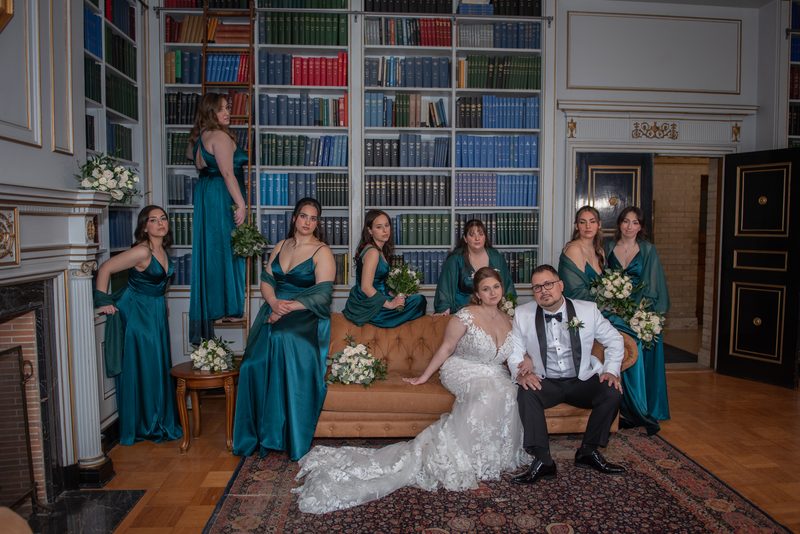 Emily and Ramon are seated on a tufted sofa in a grand library, with bridesmaids in teal dresses surrounding them.