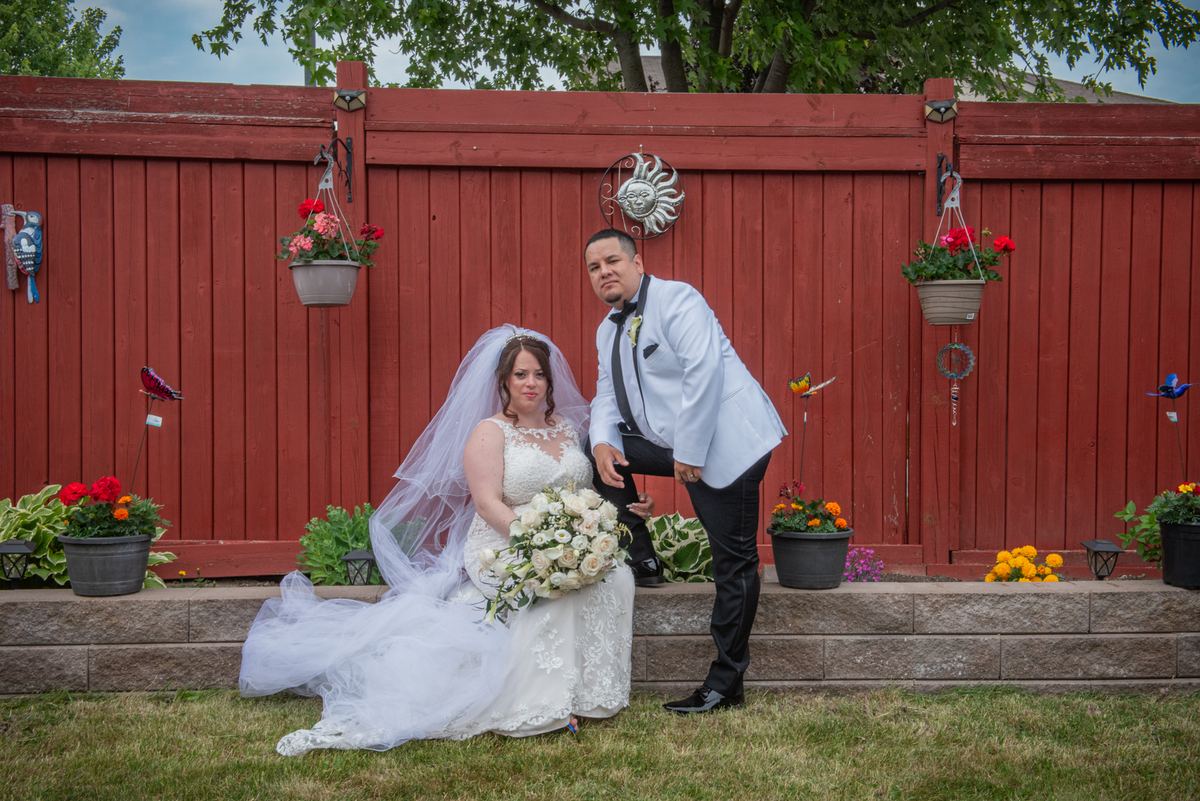 Bride sits on stone ledge with groom standing beside her in backyard with red fence and hanging flowers.