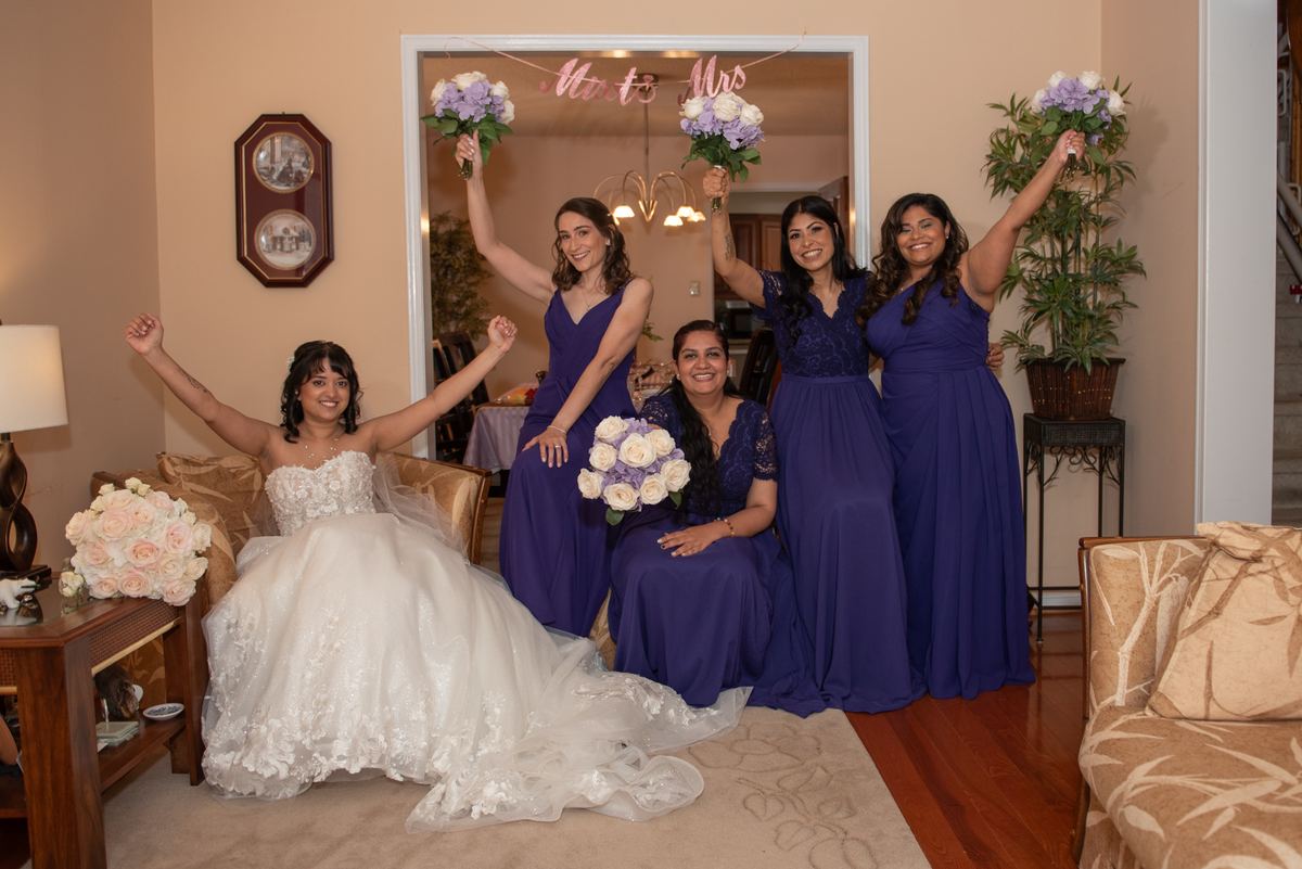 A bride and her bridesmaids celebrating indoors with raised flower bouquets, a warm glow from the lighting.