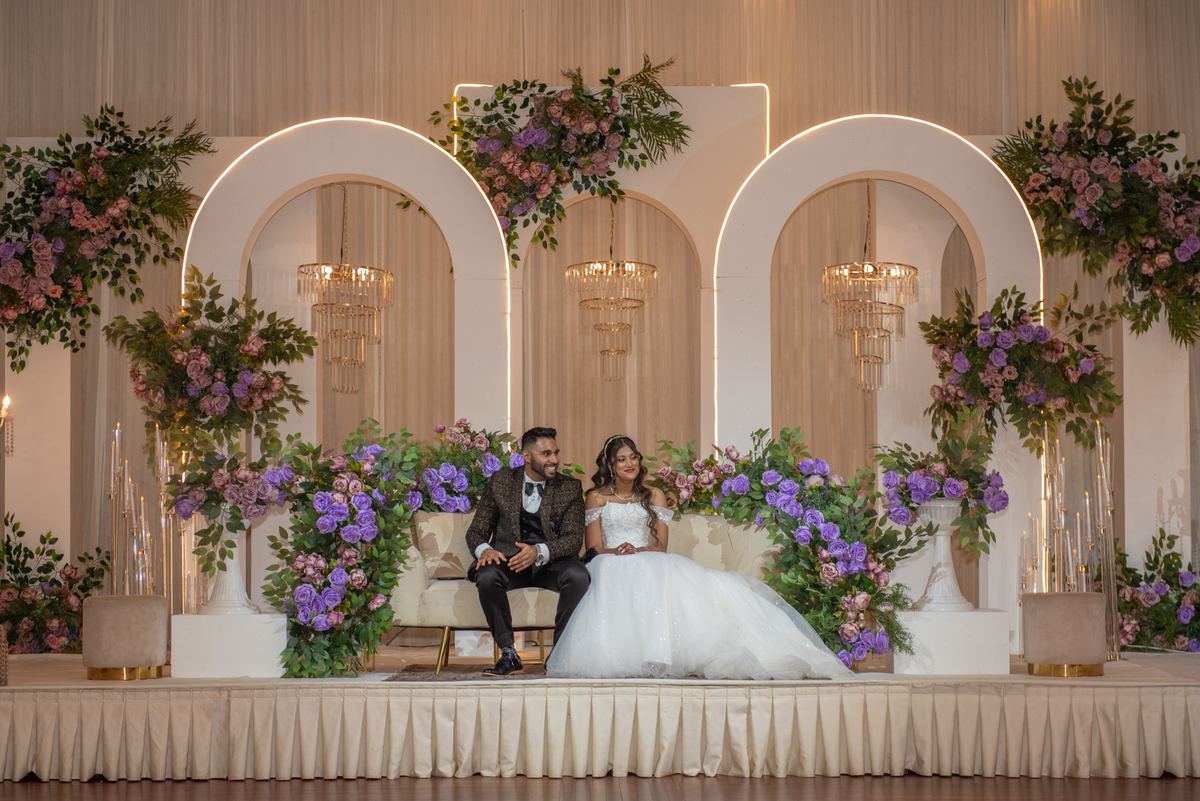 A newlywed couple sits on a decorated stage under arched backdrops with purple and pink flowers, illuminated by chandeliers.