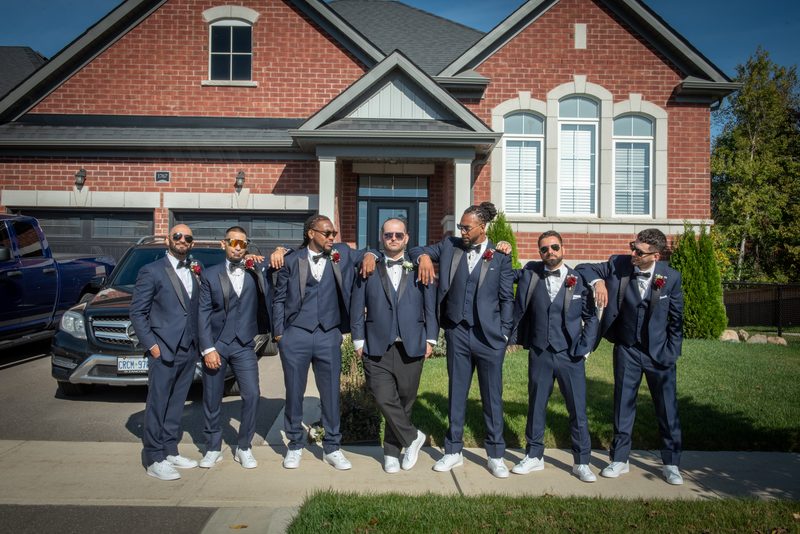 Seven men in navy tuxedos pose with arms around each other's shoulders on a sunny driveway, forming a close-knit group.