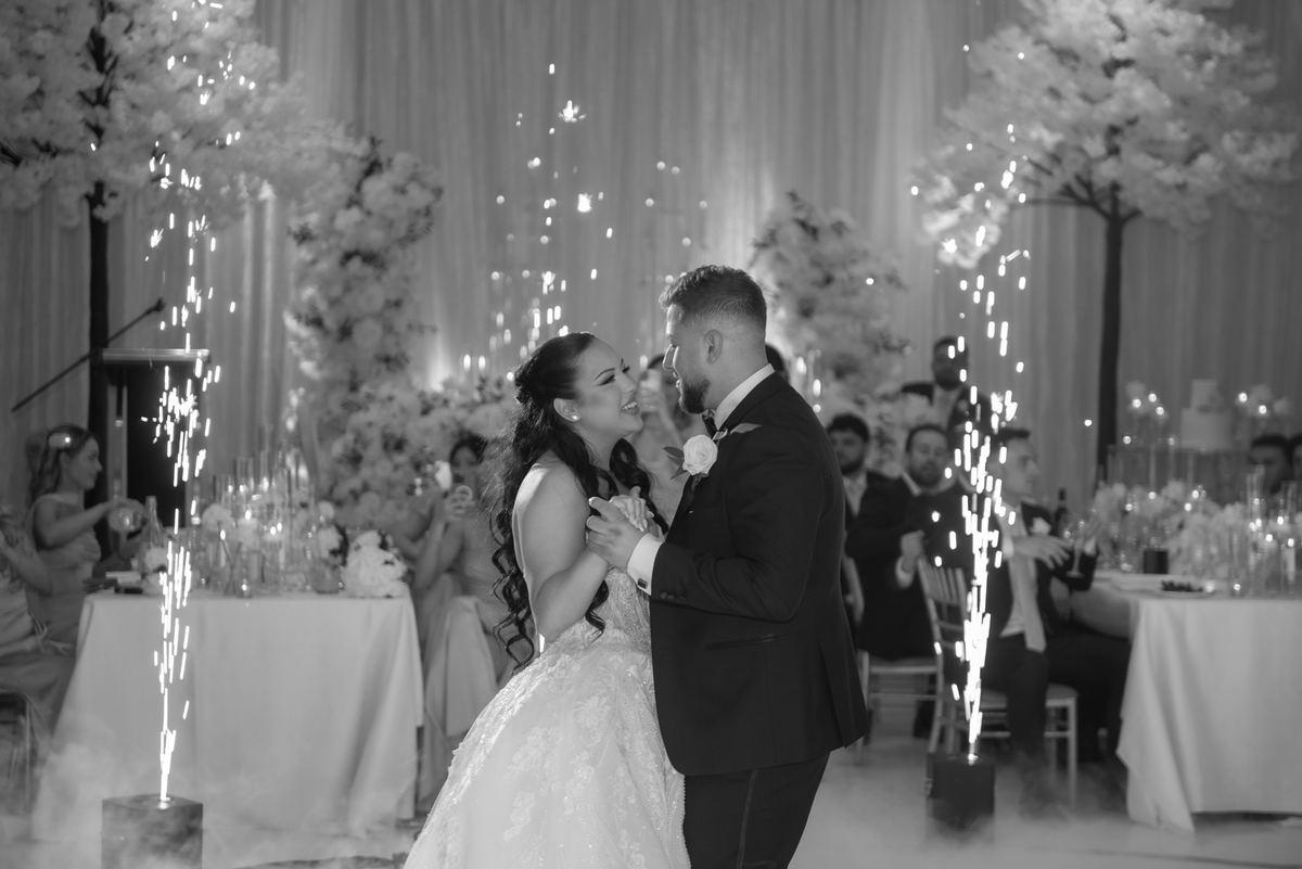 Bride and groom dance joyfully, surrounded by sparkling lights and smoke, with guests smiling in the background.