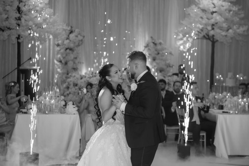 Bride and groom dance joyfully, surrounded by sparkling lights and smoke, with guests smiling in the background.