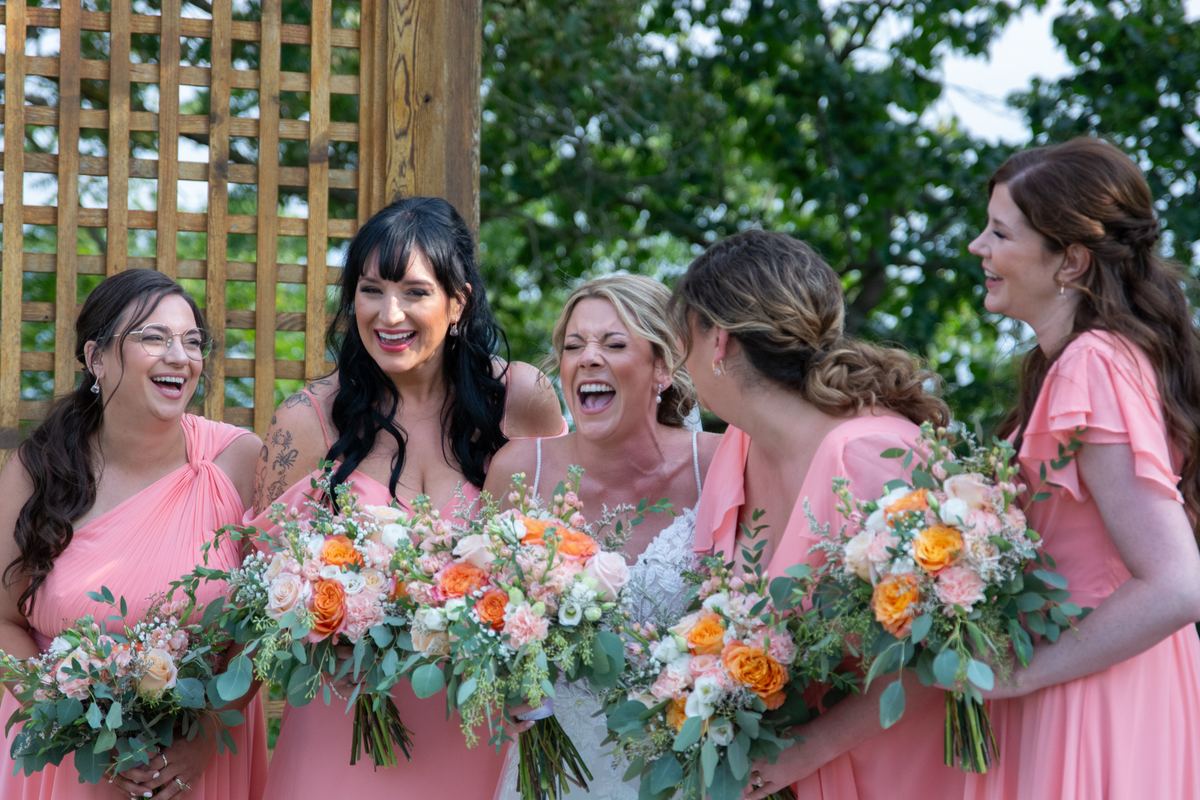 Bride and bridesmaids laugh joyfully outdoors with bouquets of colorful flowers, framed by wooden lattice.