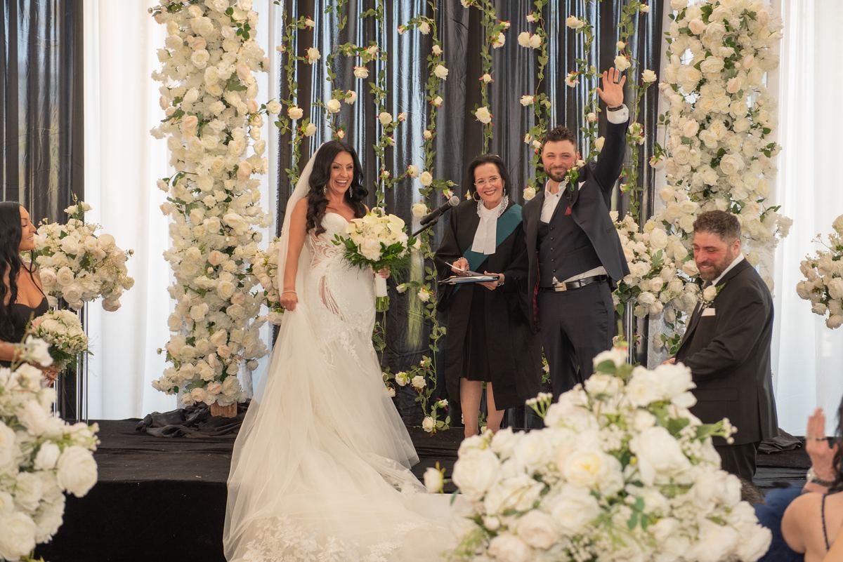 Bride in white gown and bouquet stands beside groom waving to guests at decorated stage during ceremony.